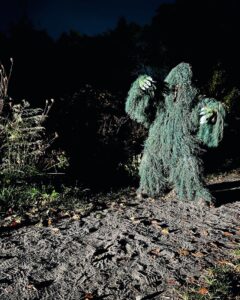 A 'monster' figure in a green ghillie suit against a dark background