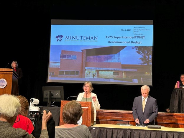 A woman speaks at a dias and a man in a suit is standing nearby. The Minuteman budget presentation is up on the screen behind her and the Town Moderator is on the stage.