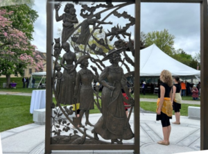 The bronze sculpture, Something is Being Done, shows several women marching down an arc. The largest figure is a woman wearing a long dress, large hat, and carrying a scroll in one hand.