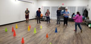 A group of people are standing in a gym. There are colored cones on the floor and a woman in a pink jacket brings a ball over to the clients.