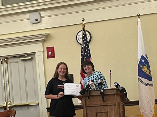 A woman stands at the podium with a younger woman. They are holding a (different) certificate.