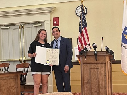 Another man stands with the young woman in front of the podium. She is holding a large certificate and smiling.