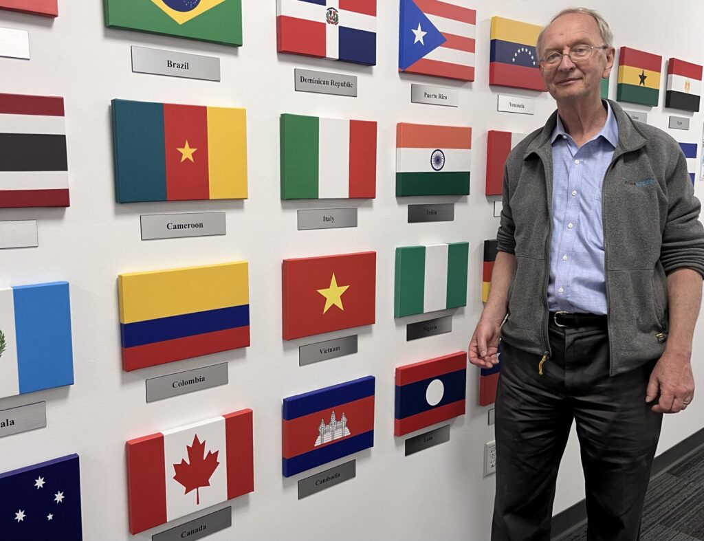 A tall man standing in front of a wall full of flags images. You can see Camaroon, Brazil, India, Canada, and many others.