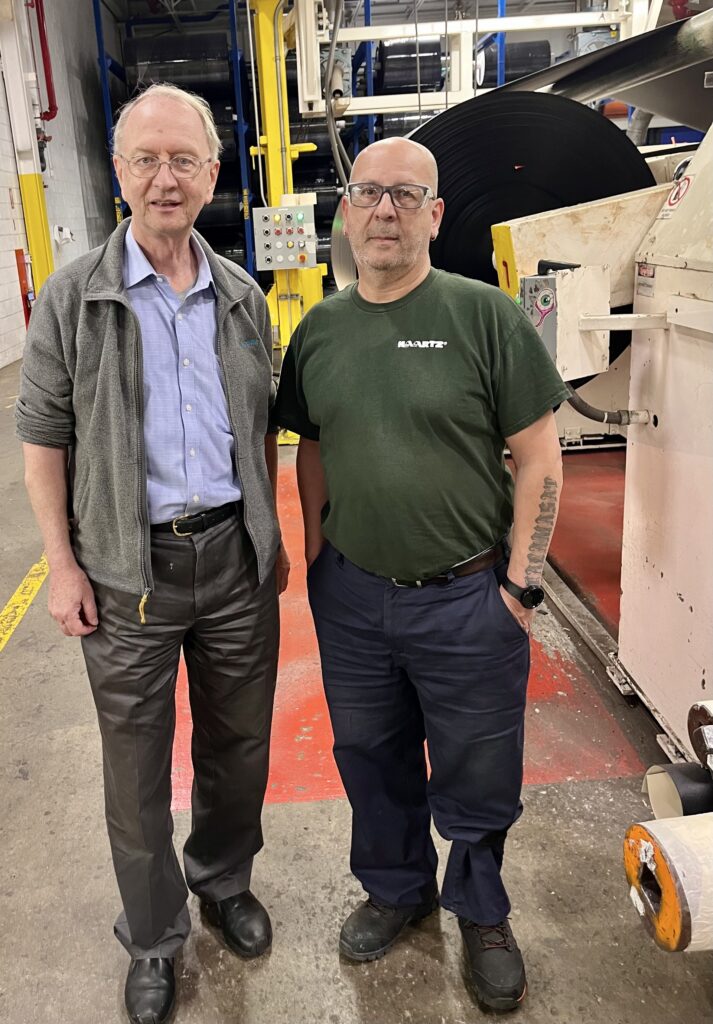 Two men, standing in front of a giant fabric roller. The man on the left is tall with grey hair and wireframe glasses, the man in the right is shorter, bald, and wearing a Haartz t-shirt.