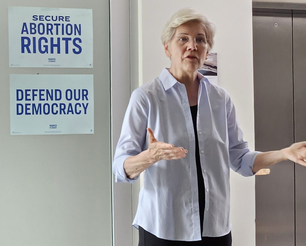 A casually-dressed woman with short blonde hair and glasses speaks in front of a wall. Behind her are Warren campaign signs that say "Secure abortion Rights" and "Defend Our Democracy."