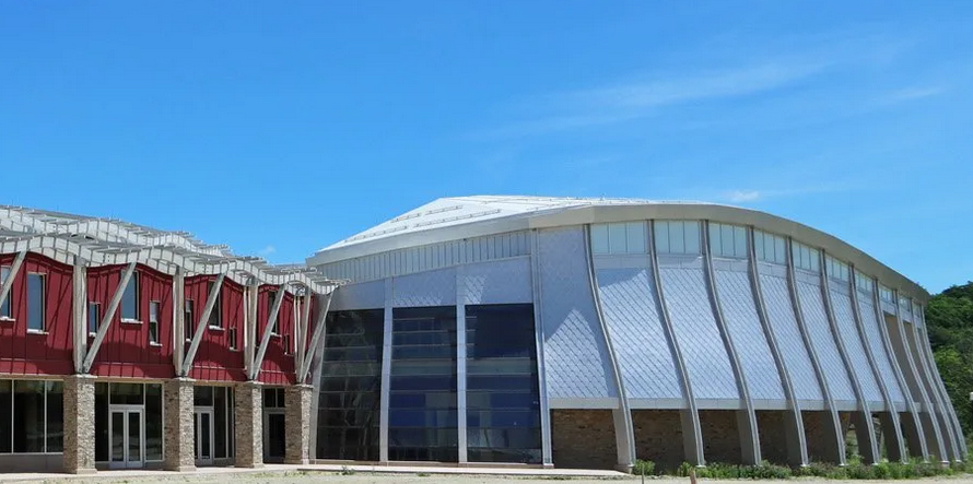 A modern building with a blue sky background. on the left side, a long building with stone columns on the first floor, red siding on the second floor and a modern multi-part awning. On the right, a silvery building soars. It has a white curved roof and silvery panels between concrete pillars.