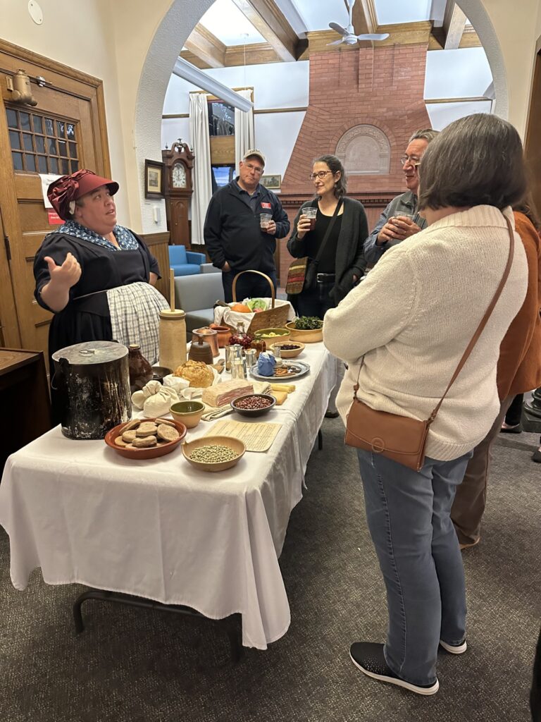 A woman wearing a colonial-style dress stands in front of a table covered with foods and implements from a colonial kitchen including a small butter churn, several pots, and a basket of small, English-muffin type breads.