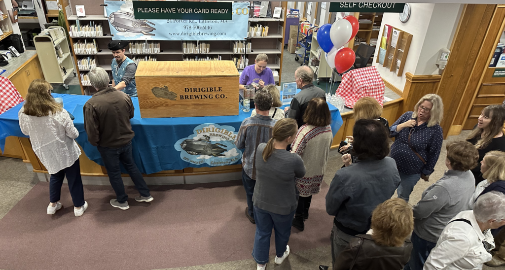 The AML checkout counter sports a blue Dirigible Brewing tablecloth and a large wooden box that hold the kegs. There are people standing in line and two people serving up brews behind the counter. The photo is taken from the stairs, so you see the tops of people's heads.
