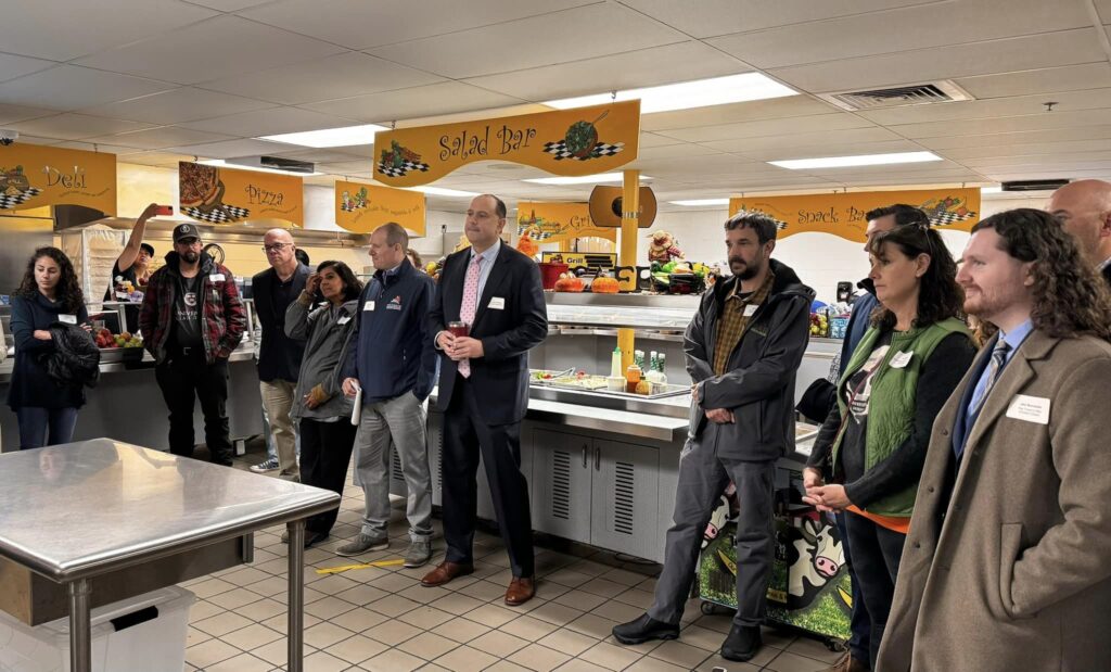 A group of people stand in front of the salad bar in a cafetiera. They are listening to someone off-camera.