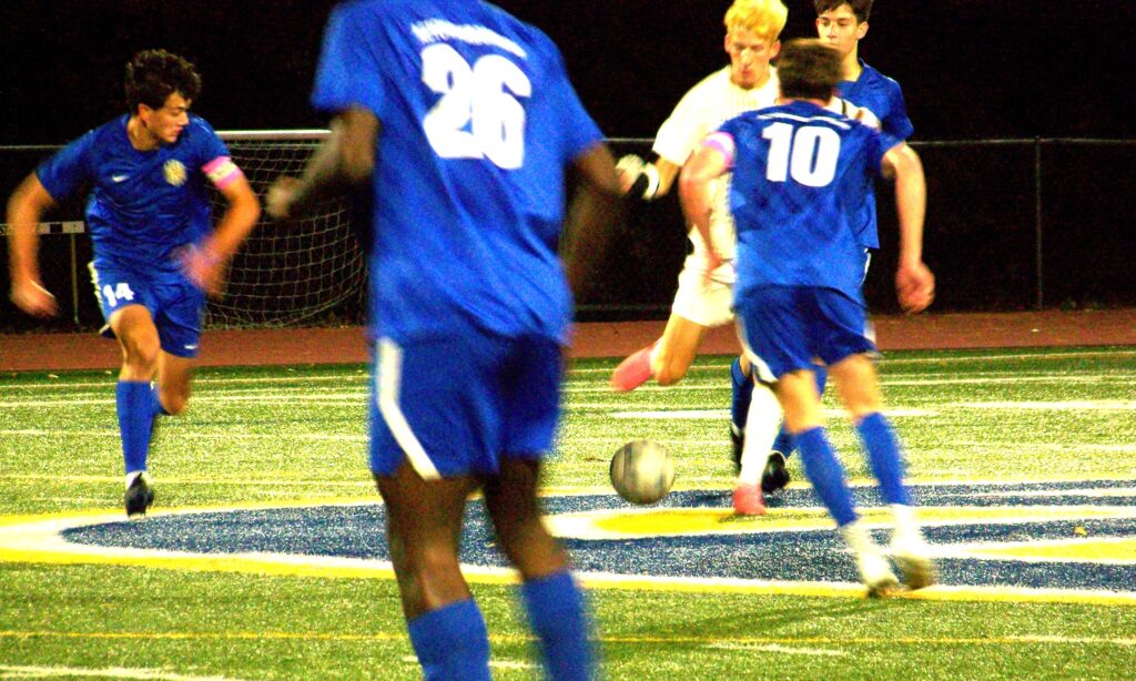 Young men (mostly with Revs jersys, run towards a ball that a man in an Algonquin jersey is about to kick.