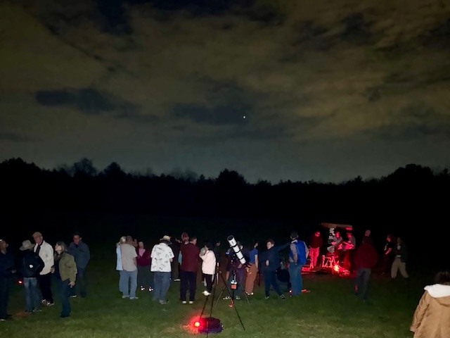 On a cloudy-ish night a group of people are gathered. You can see a large telescope in the foreground and some red lights scattered around.