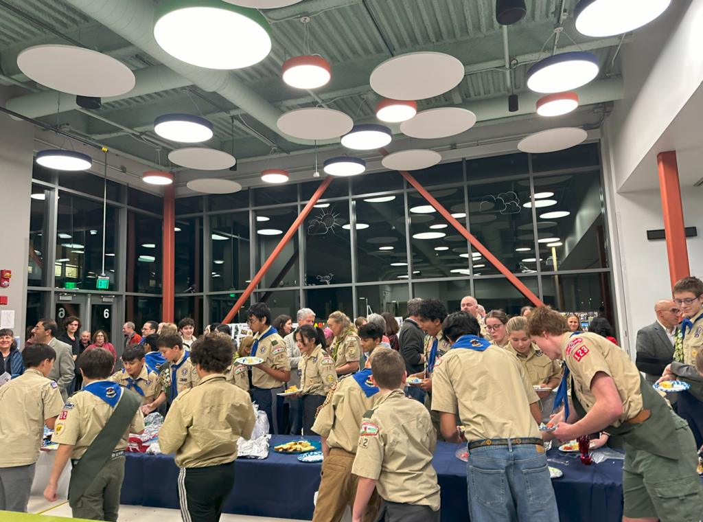 A crowd of scouts of various ages fill plates at tables full of food in the Boardwalk Campus cafeteria.