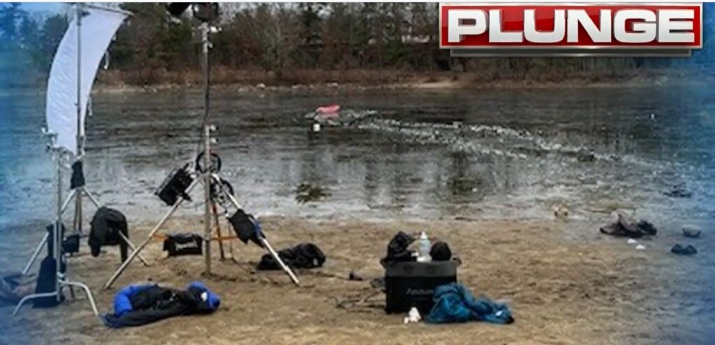 A red kayak bobbles in the water, there's a broken path in the ice between the kayak and the shore. On the shore, a bunch of film equipment stands idle.
