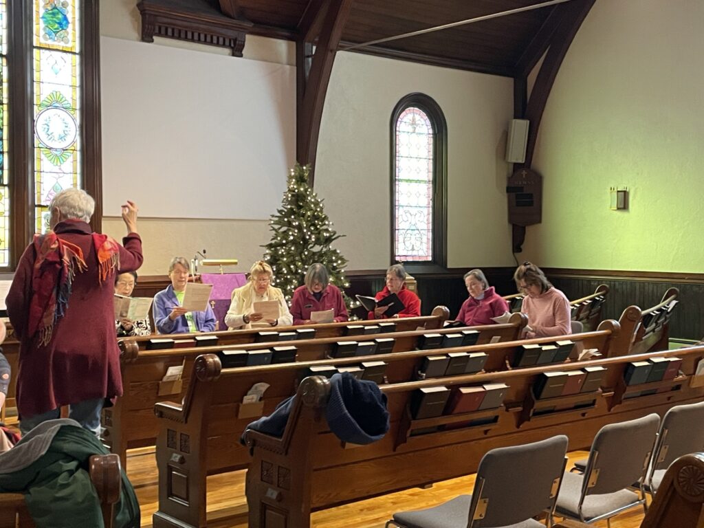 A group of women are seated in the church sanctuary. They are holding music and singing. On the right, an older woman is leading the choir. Coats and bags rest on the pews.