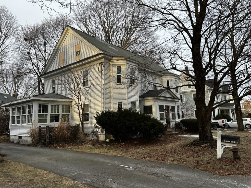 A white house. In the eaves, windows appear to be covered in plywood.