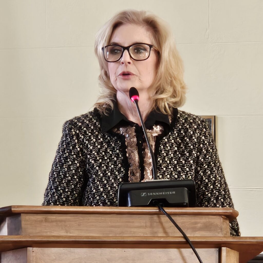 A woman with blonde hair and glasses speaks at a podium.