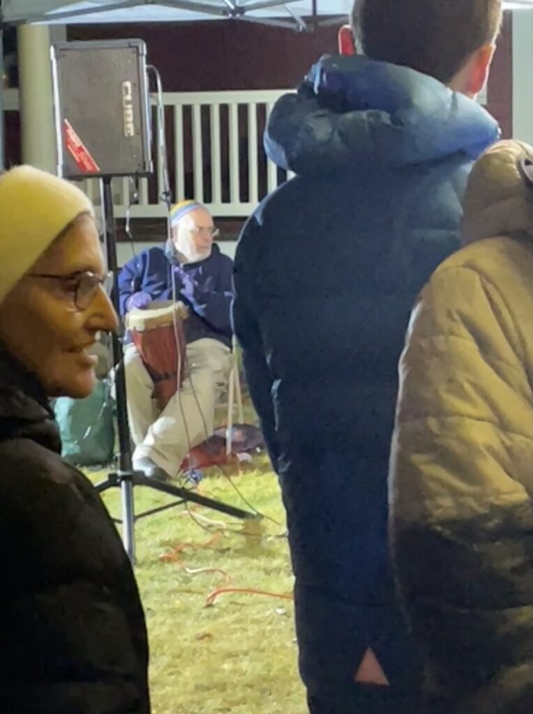A man is seated in front of the Red House playing a wood drum. People are milling around in front of him.