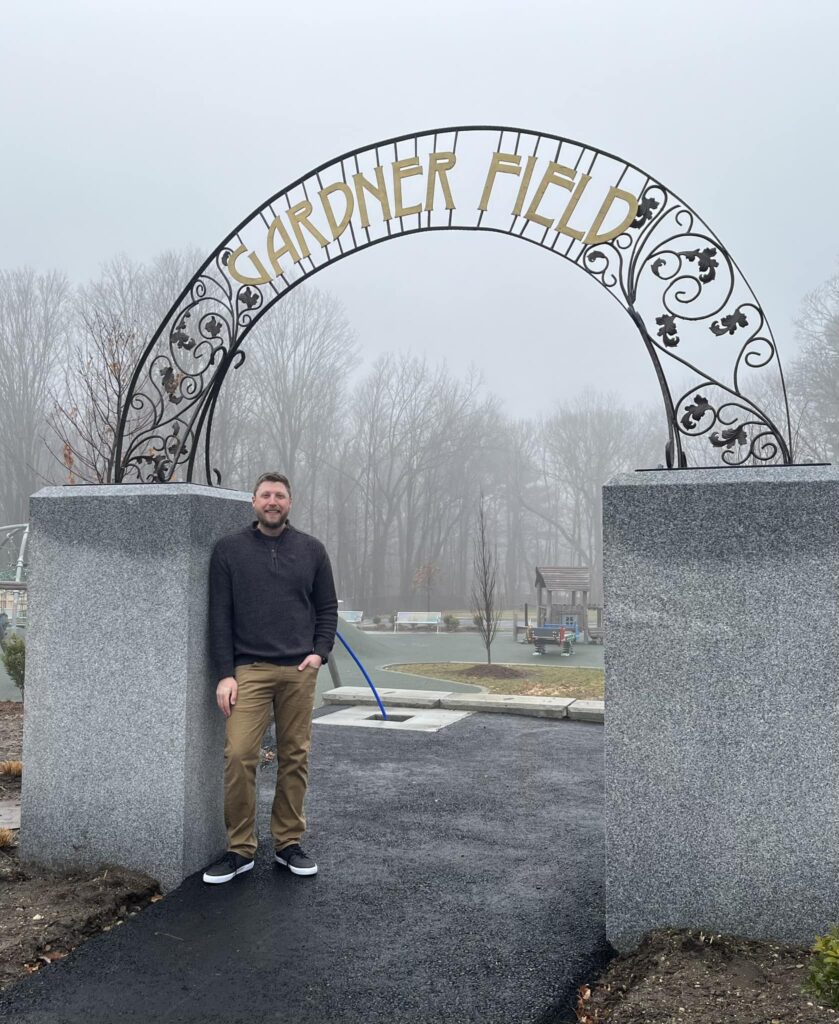 A man leans against a concret pillar that holds the Gardner Field arch. The arch has black vines with leaves and says Gardner Field in large brass letters.