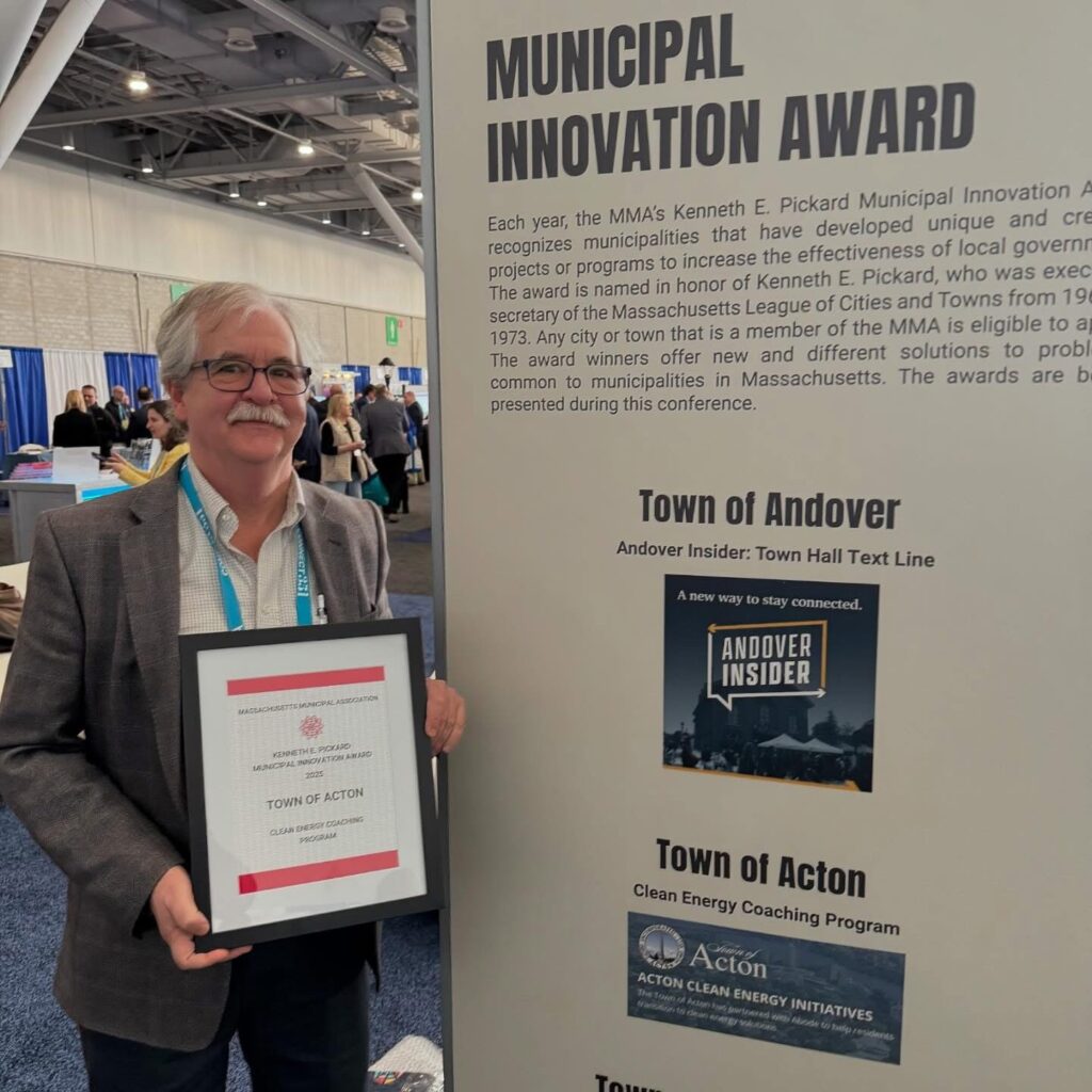A man with grey hair and mustache holds an award to the town of Acton. He stands next to a large poster that says Municipal Innovation Award and highlights both Andover and Acton.