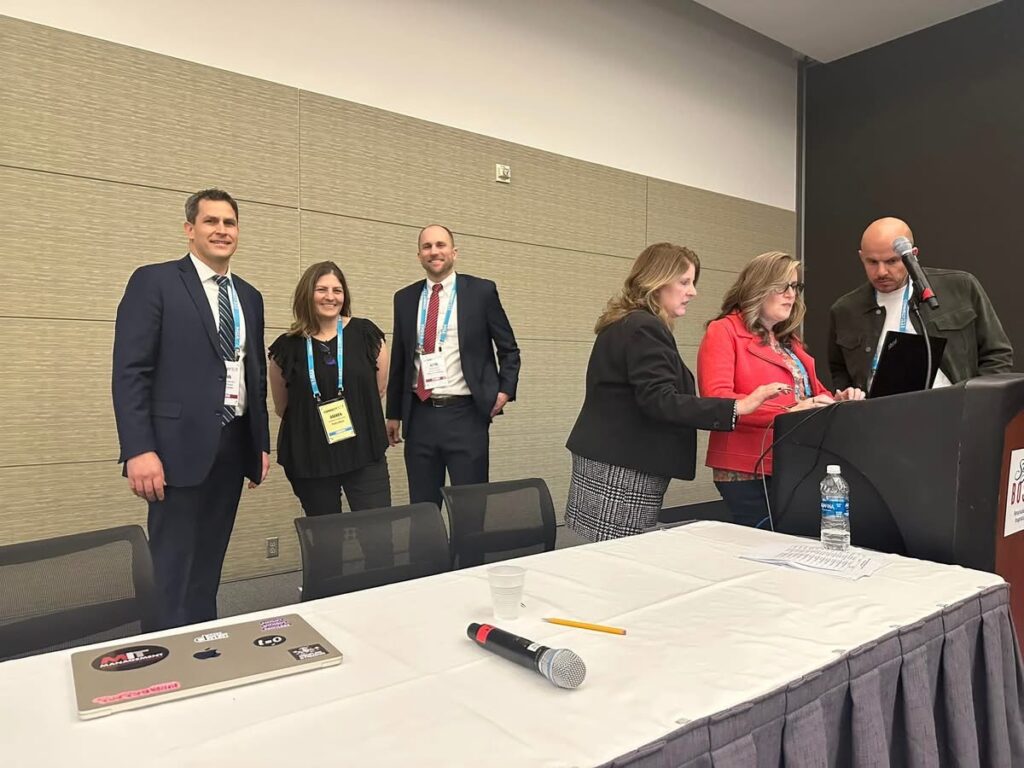 Two groups of people in a room. One group stands behind a table, another group is at a podium.