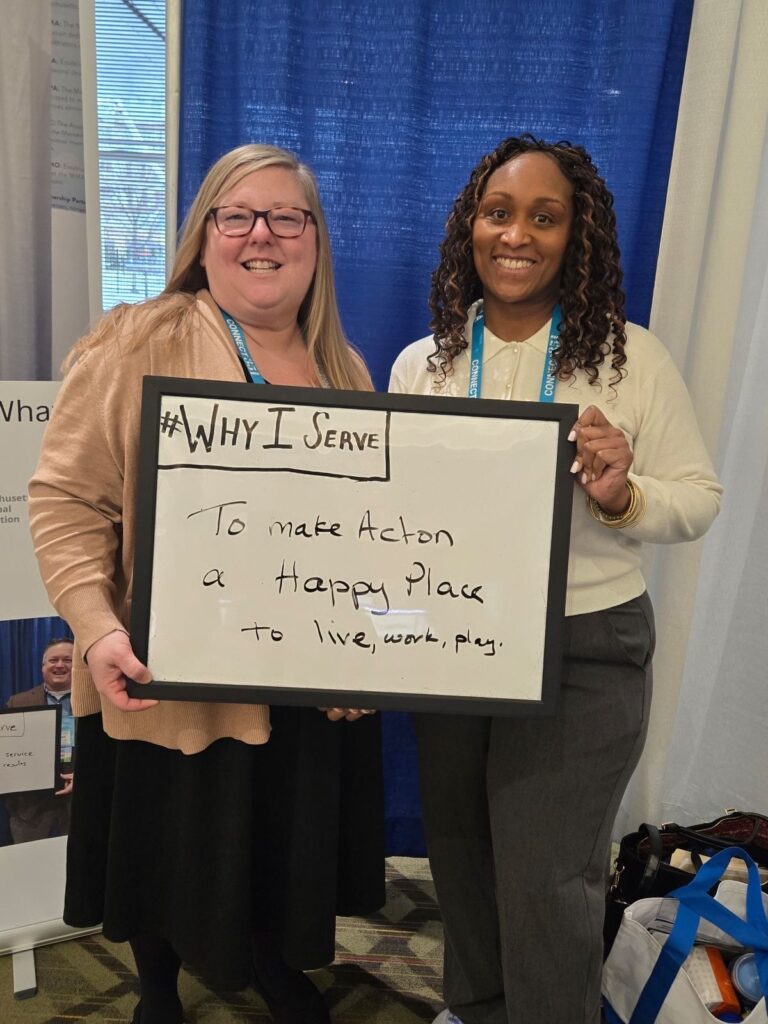Two women, one blonde, one curly black hair hold a white board that says #Why I serve in the corner. Their response is "To make Acton a happy place to live, work, play."