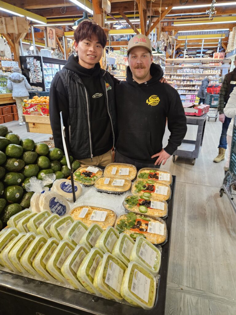 Two men stand before an array of dips, tortillas, and a tall stack of avocados.
