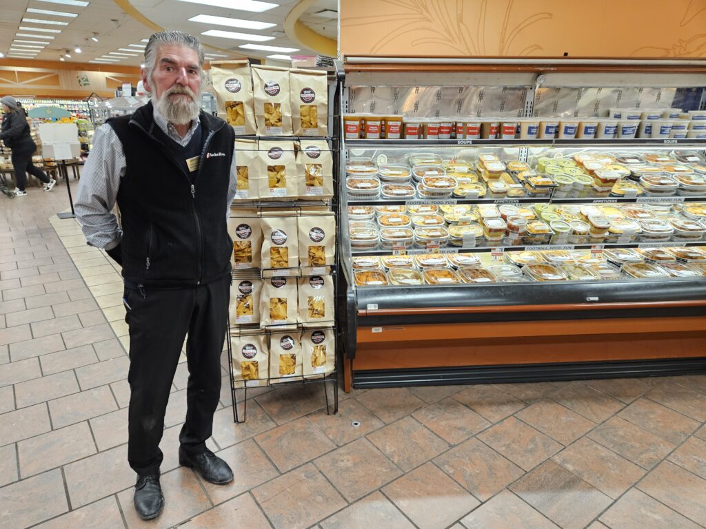 A tall bearded man stands next to a display with chips and a refrigerator case full of dips and snacks.