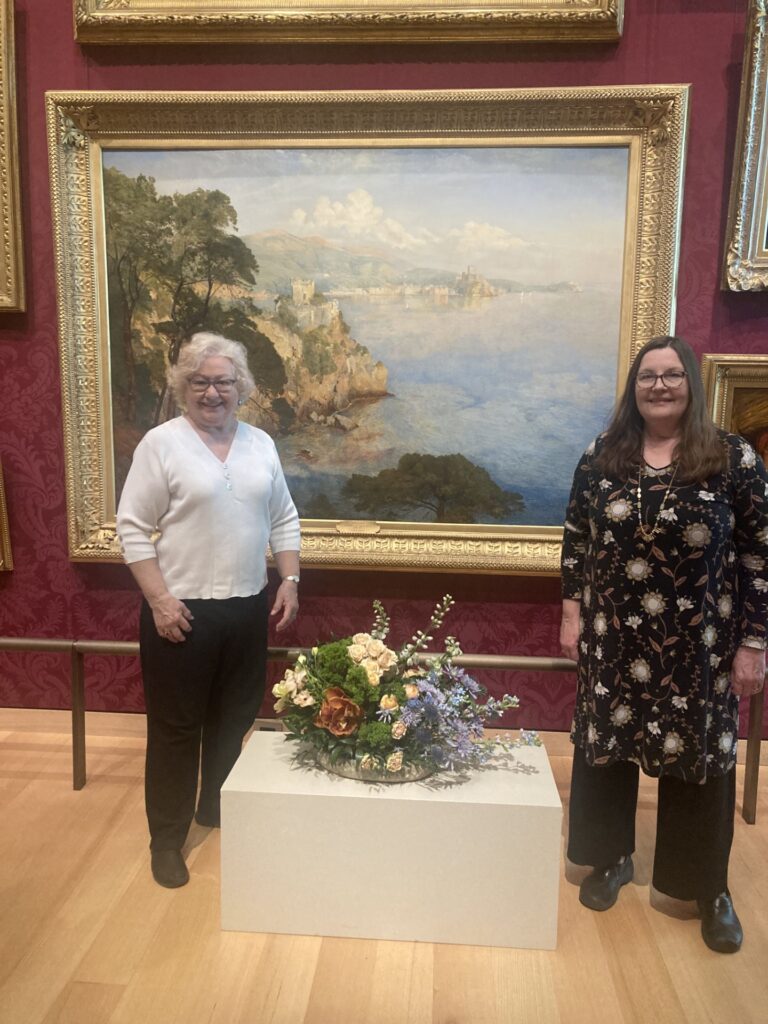Two smiling women stand prudly in front of their lovely arrangement.