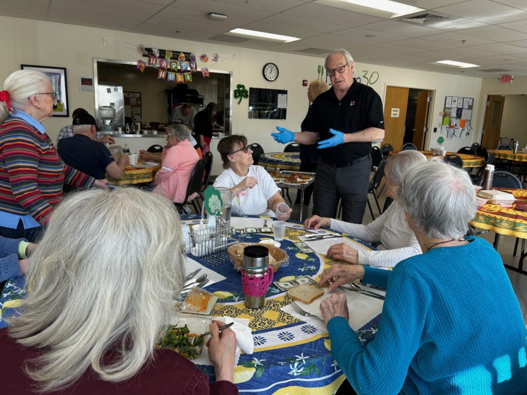 Five mostly grey-haired diners around a round table listen to a grey-haired man with blue latex gloves talking. The diners have bread or salad in front of them and the man has a tray of food he is about to deliver to the table.