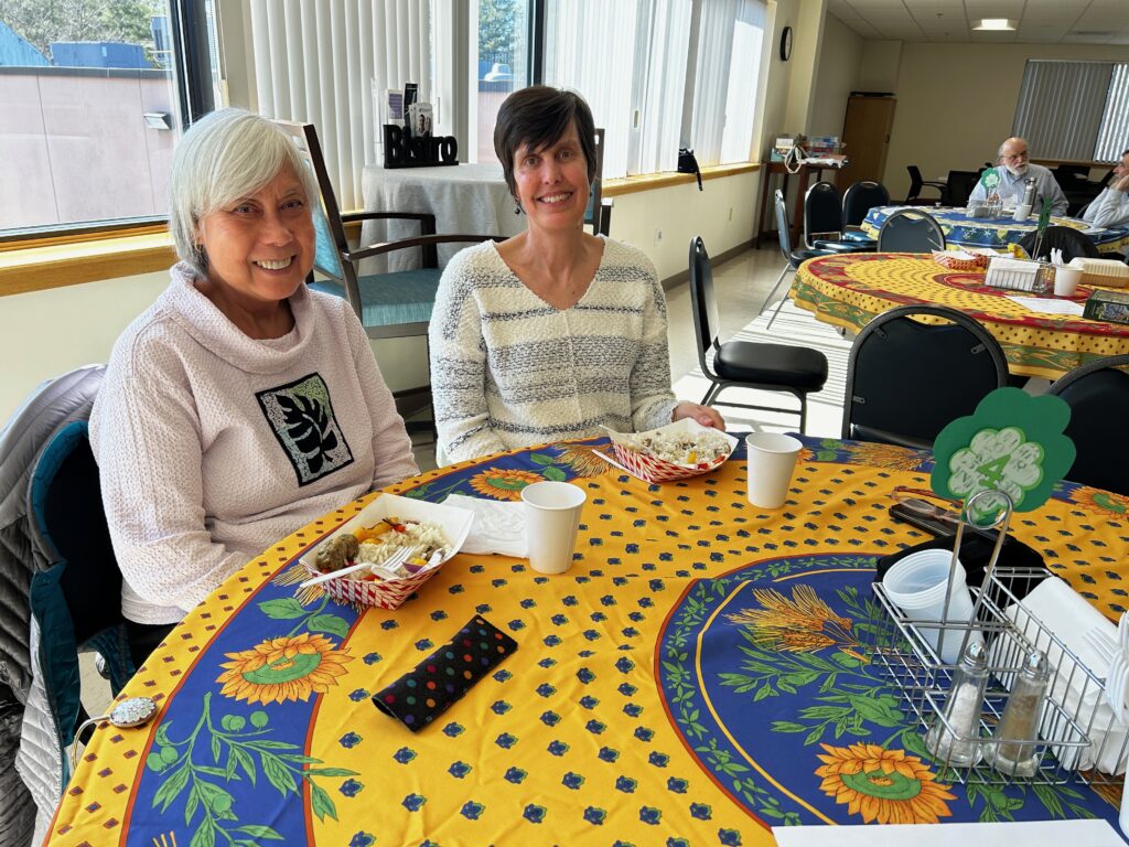 Two women smile as they sit across a table covered with a yellow and blue patterned tablecloth in front of cardboard bowls of food. Other tables are beyond, one with two men talking. Large windows with vertical blinds at the left.