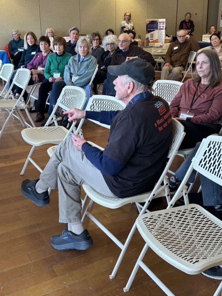A group of people are sitting in chairs. The man at the front chair, wearing a 350Mass T-shirt, is speaking.