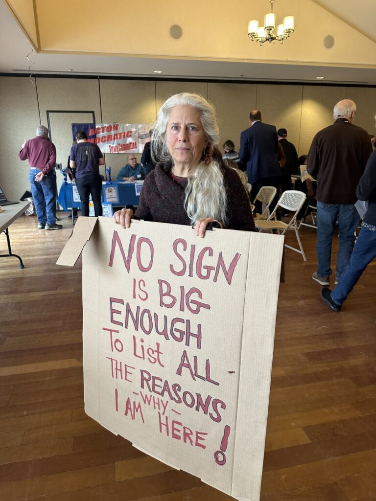 A woman with long white hair holds a sign that says: "No Sign is Big Enough to List All the Reasons why I Am Here!"