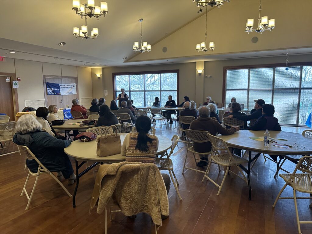 People sit at round tables in a hall. At the very back, a woman stands at a podium.