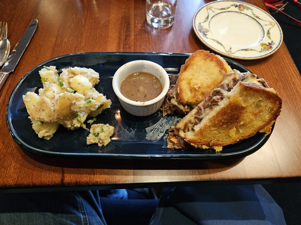 An elegant lunch: Potatoes, a ramiken of au jus, and a grilled cheese and meat sandwich, all on a dark oval plate.