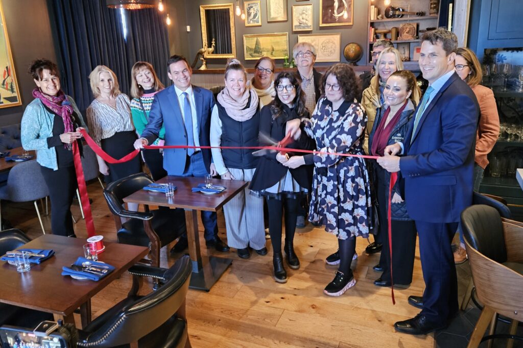 A group of people stand behind the ribbon while the owners cut the red ribbon with oversized scissors.