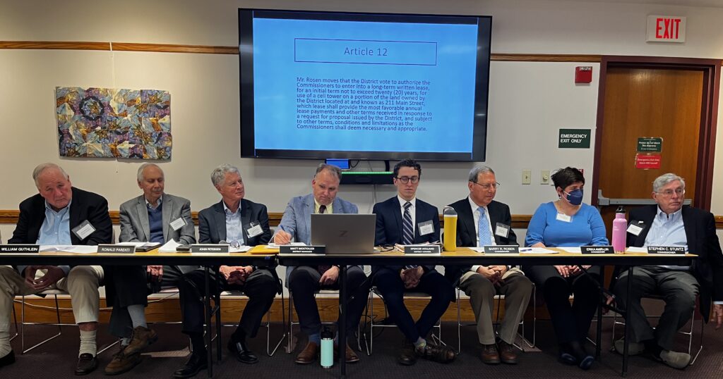Seven men and one woman, in business attire, sitting at a long table, looking offscreen to their left. Above them is a large screen displaying a block of text, headed "Article 12."
