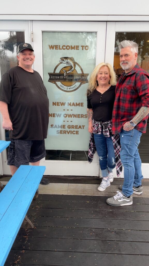 Three people, two men and a woman, stand in front of a door with an Acton Seafood Company logo (a jumping fish).