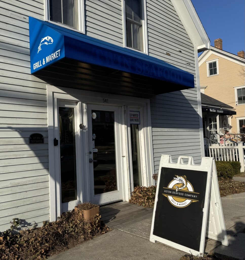 Acton Seafood Company sign on a sandwich board in front of a grey building with a blue awning.