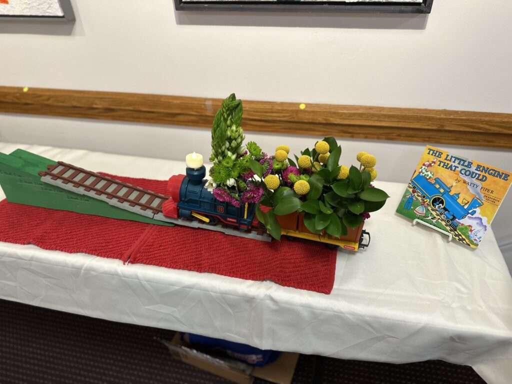 A toy train heading up a small track hauling a car with yellow and pink flowers.