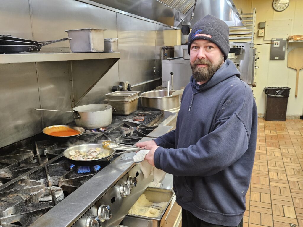 A bearded man in a blue sweatshirt and a watch cap cooks on a commercial gas stove.
