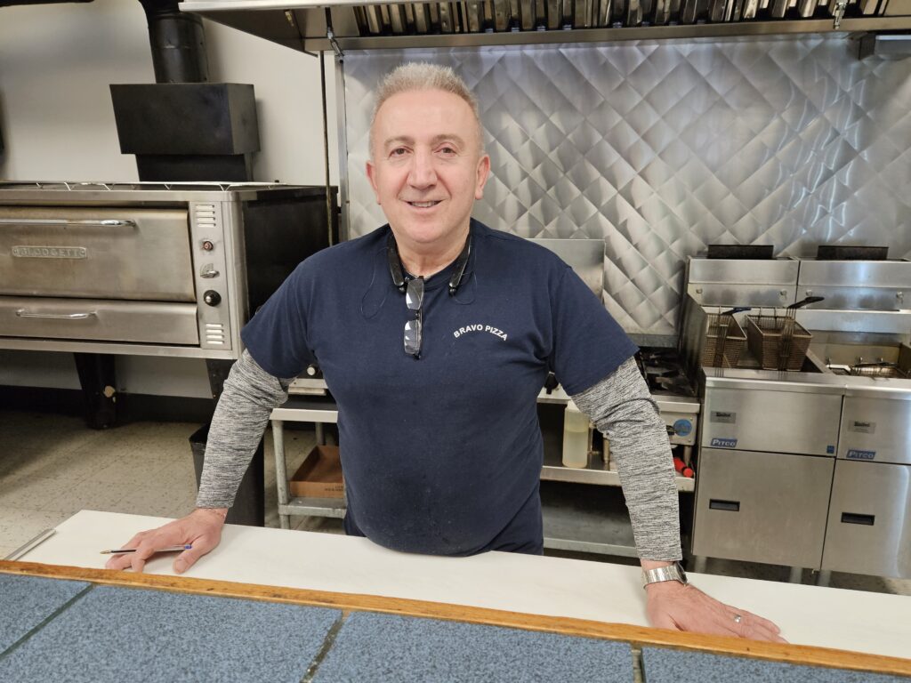 A man wearing a Bravo Pizza t-shirt stands at a counter. A commercial kitchen is in the background.