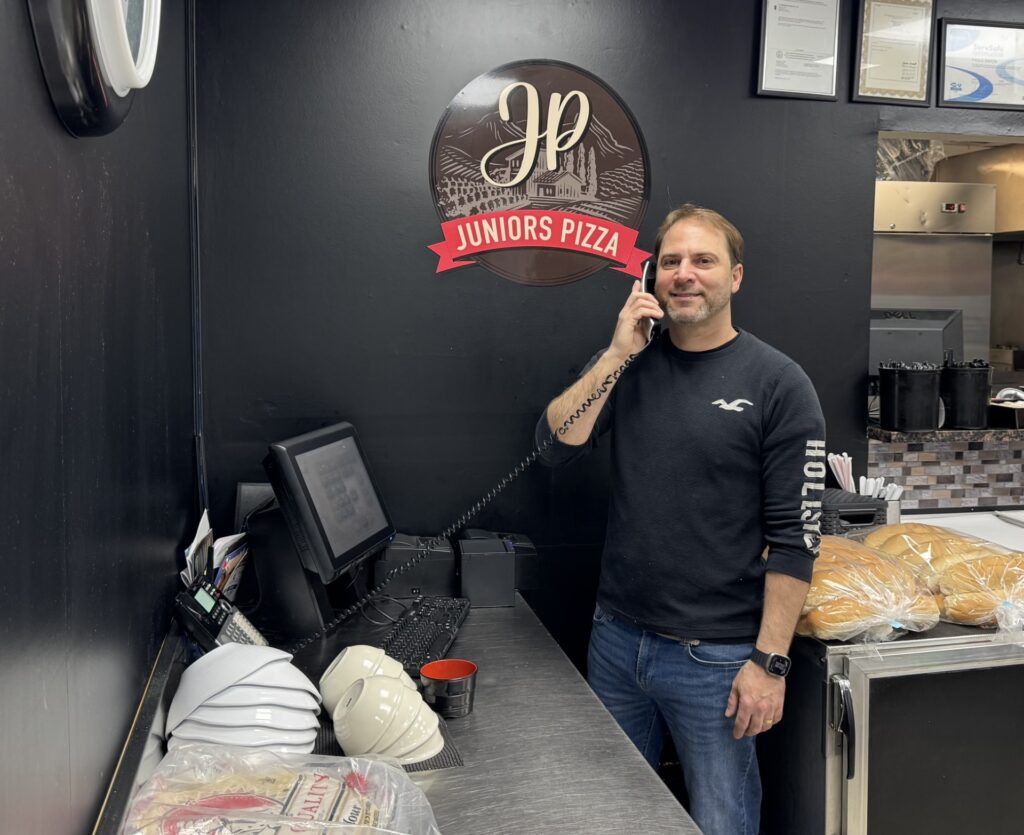 A man in a black t-shirt answers the phone in front of the Junior's Pizza sign.