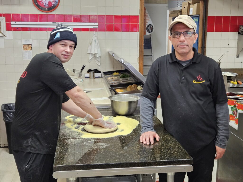 Two men in a commercial kitchen.The younger man on the right is kneading a batch of pizza dough.