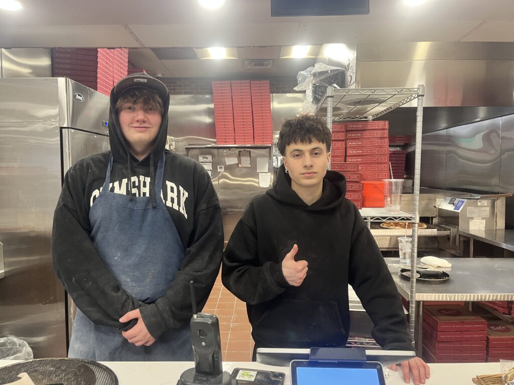 Two young men stand at the counter in front of a commercial kitchen. The young man on the right gives a thumbs up.