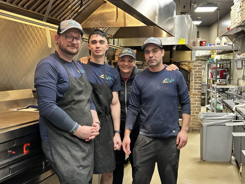 A group of four men stand in a commercial kitchen. In the back, you can see a brick oven.