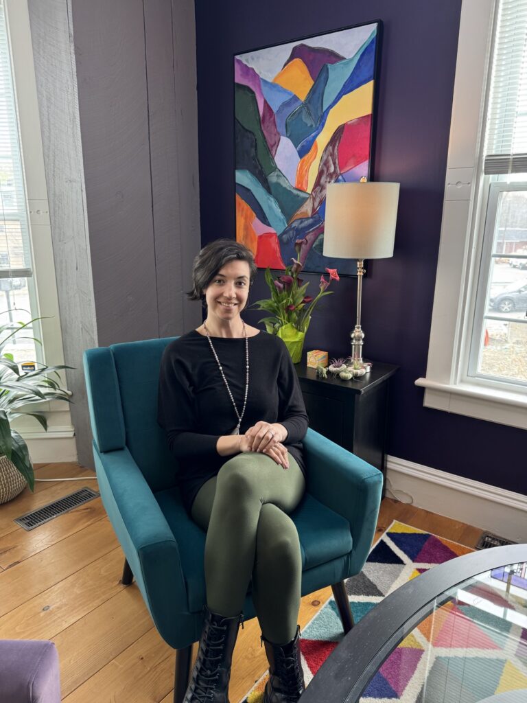 A woman with an asymmetrical haircut sits on a teal blue chair. A colorful piece of modern art is on the wall in the background.