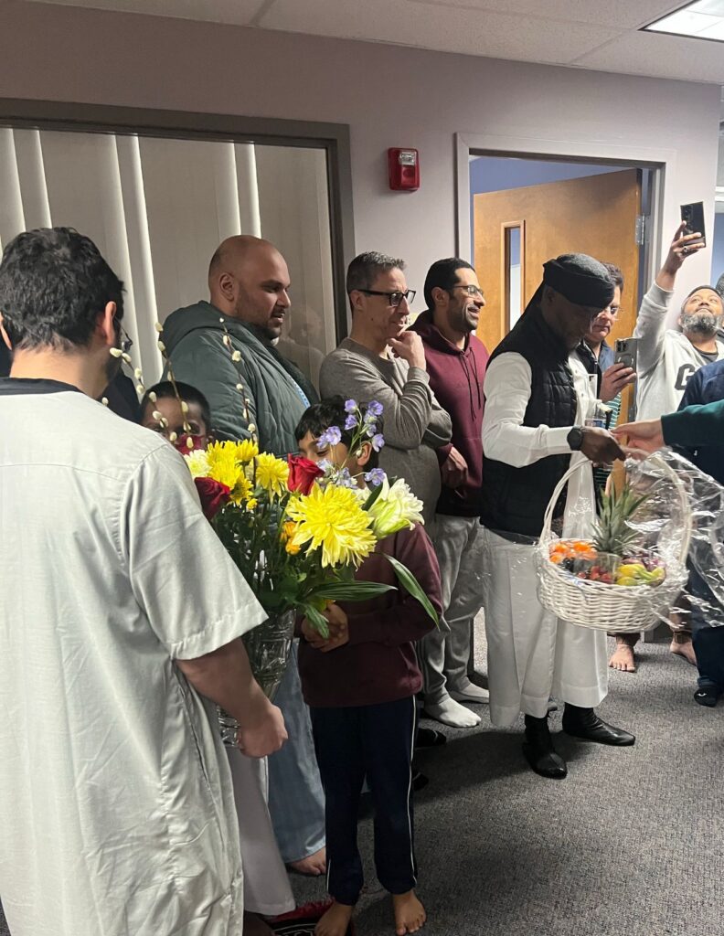 A group of men hold look on and take photographs with phones as two of them hold an arrangement of flowers and a basket of fruit.