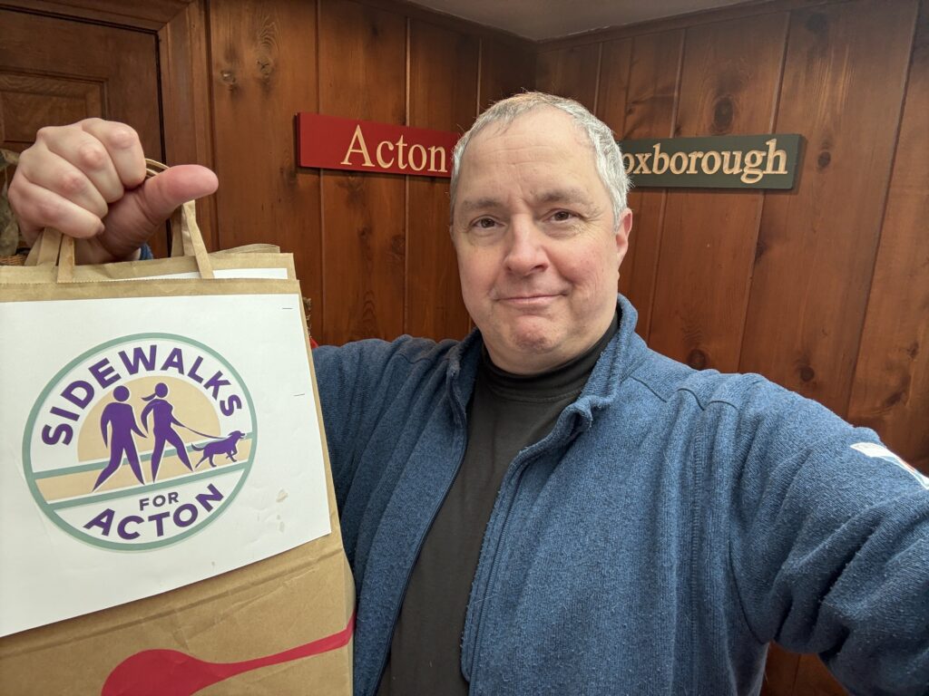 Man holds up a bag labeled "Sidewalks for Acton, with a stylized graphic of a  man, woman, and leashed dog walking on a sidewalk.