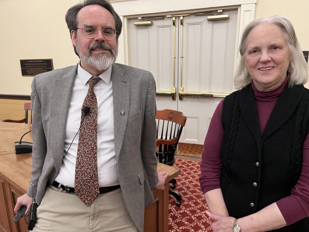 A man in a suit and tie leans on a desk next to a woman wearing a black sweater vest.