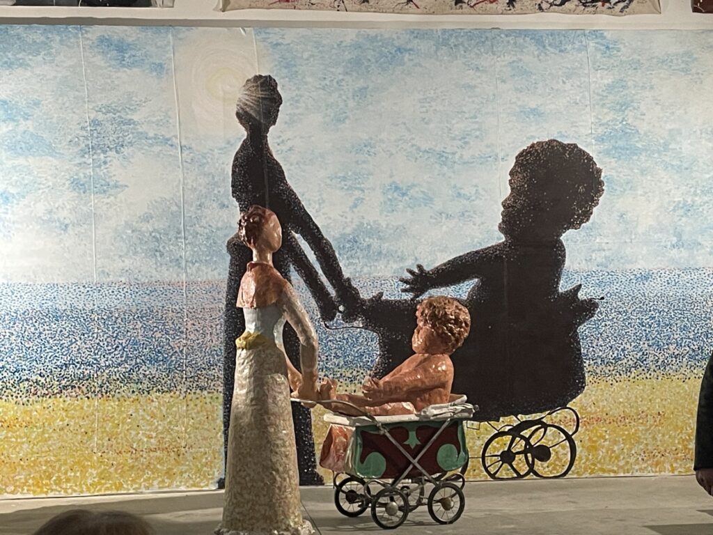 A fiberglass woman rolls and old fashioned baby carriage. In the carriage is a baby so large that it barely fits in the pram.
